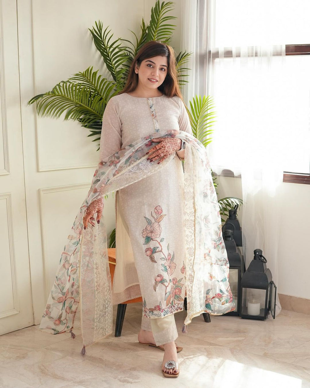 Woman in a light pink traditional outfit with floral patterns in a room with plants and decor.